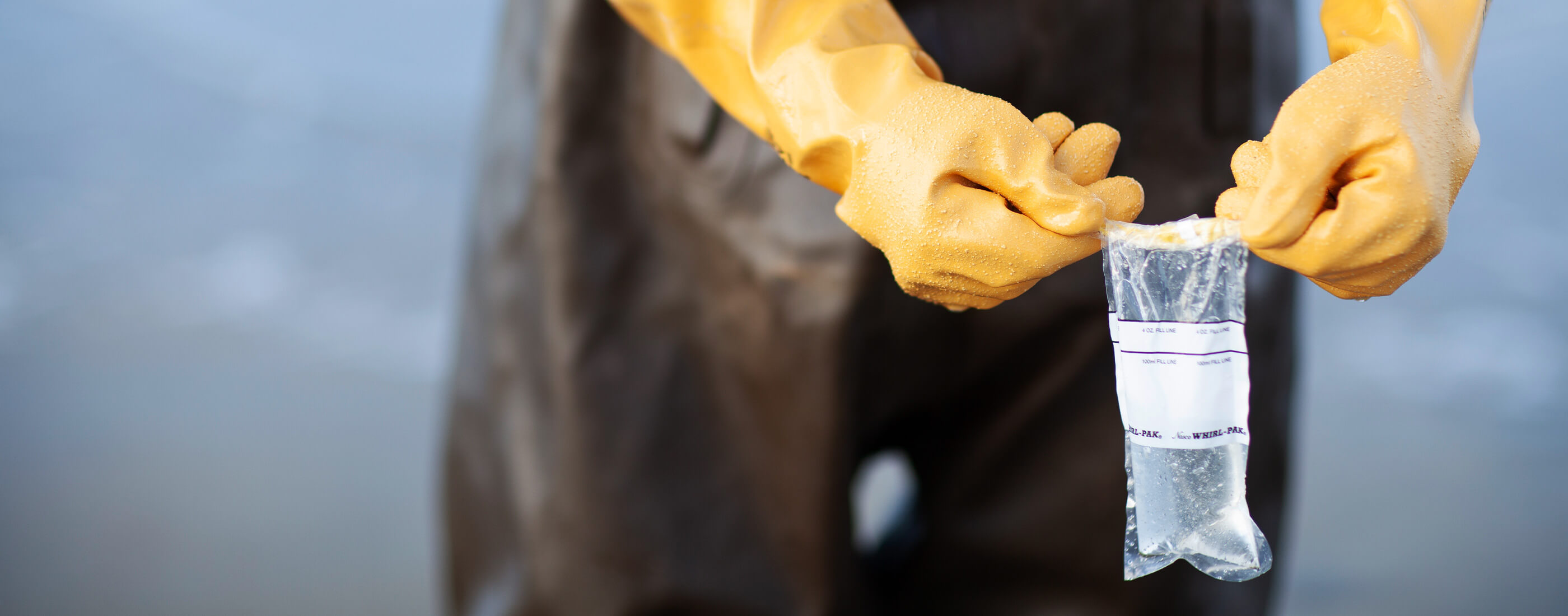 Hands in yellow gloves hold up water sample from the beach